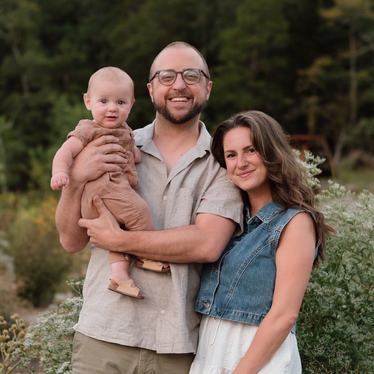 Jon holding baby Leo next to Katherine outdoors among greenery