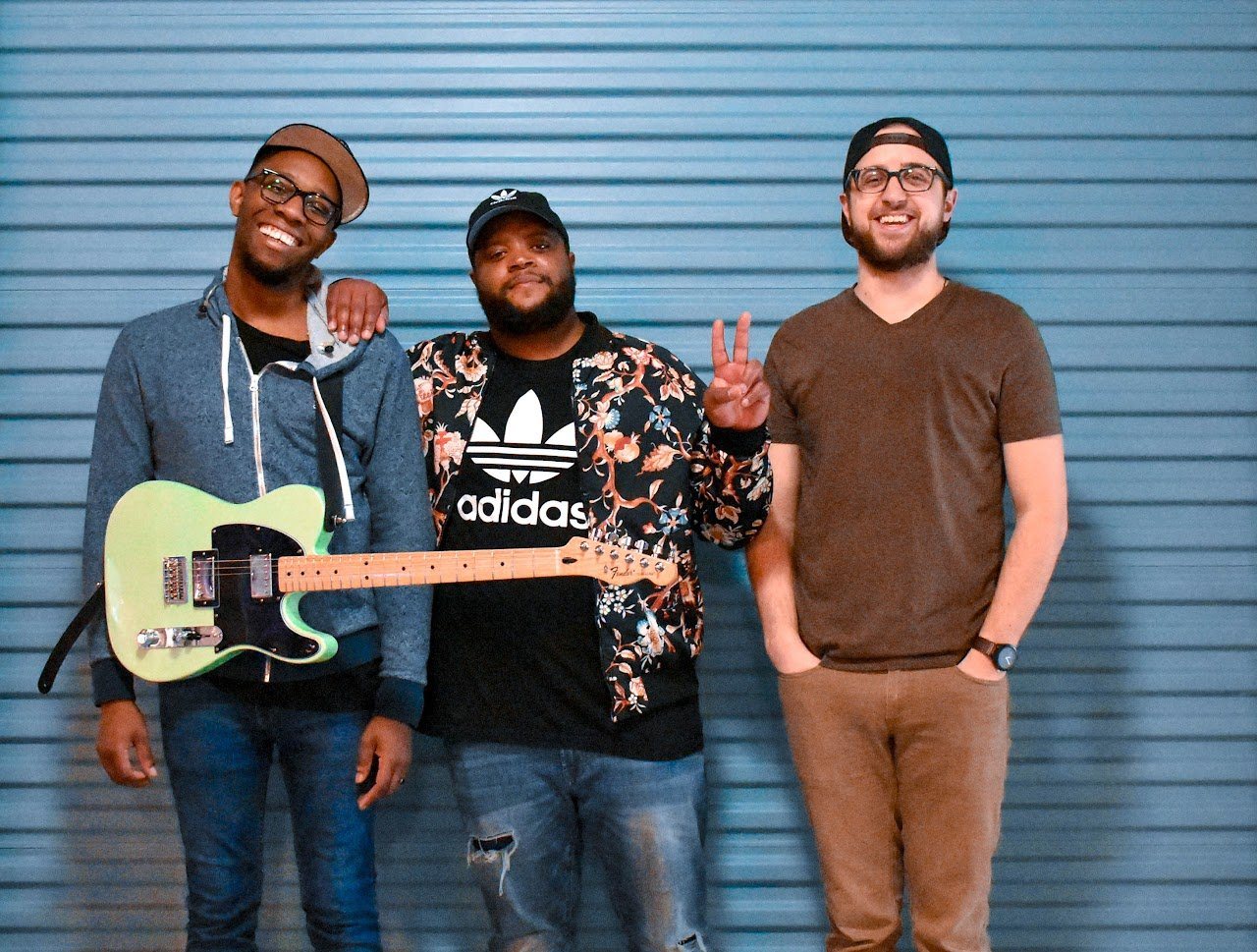Jon standing with two friends against a blue wall, one holding an electric guitar