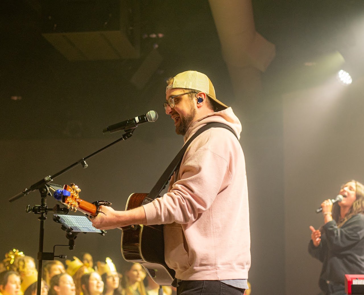 Jon leading worship on acoustic guitar, singing into a microphone under stage lights