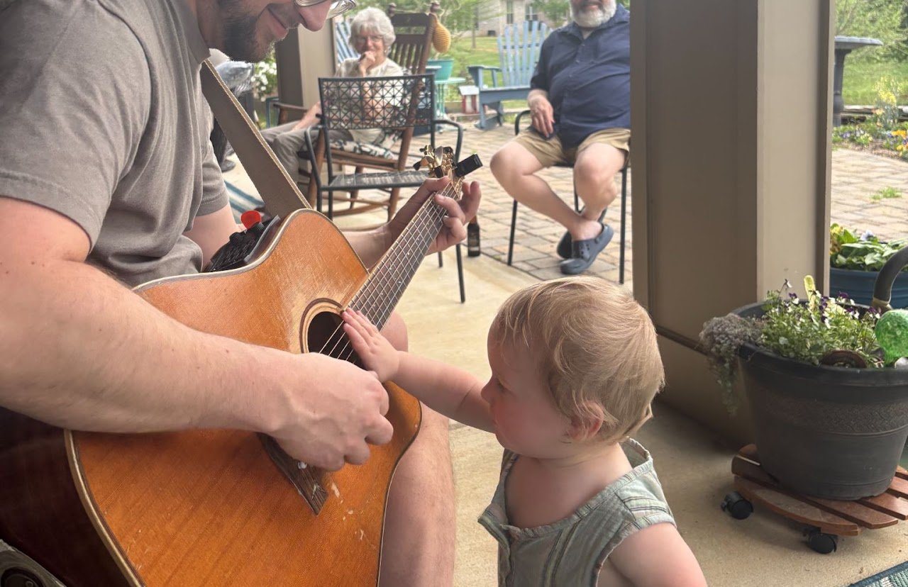 Jon playing acoustic guitar on a patio while a toddler reaches for the strings