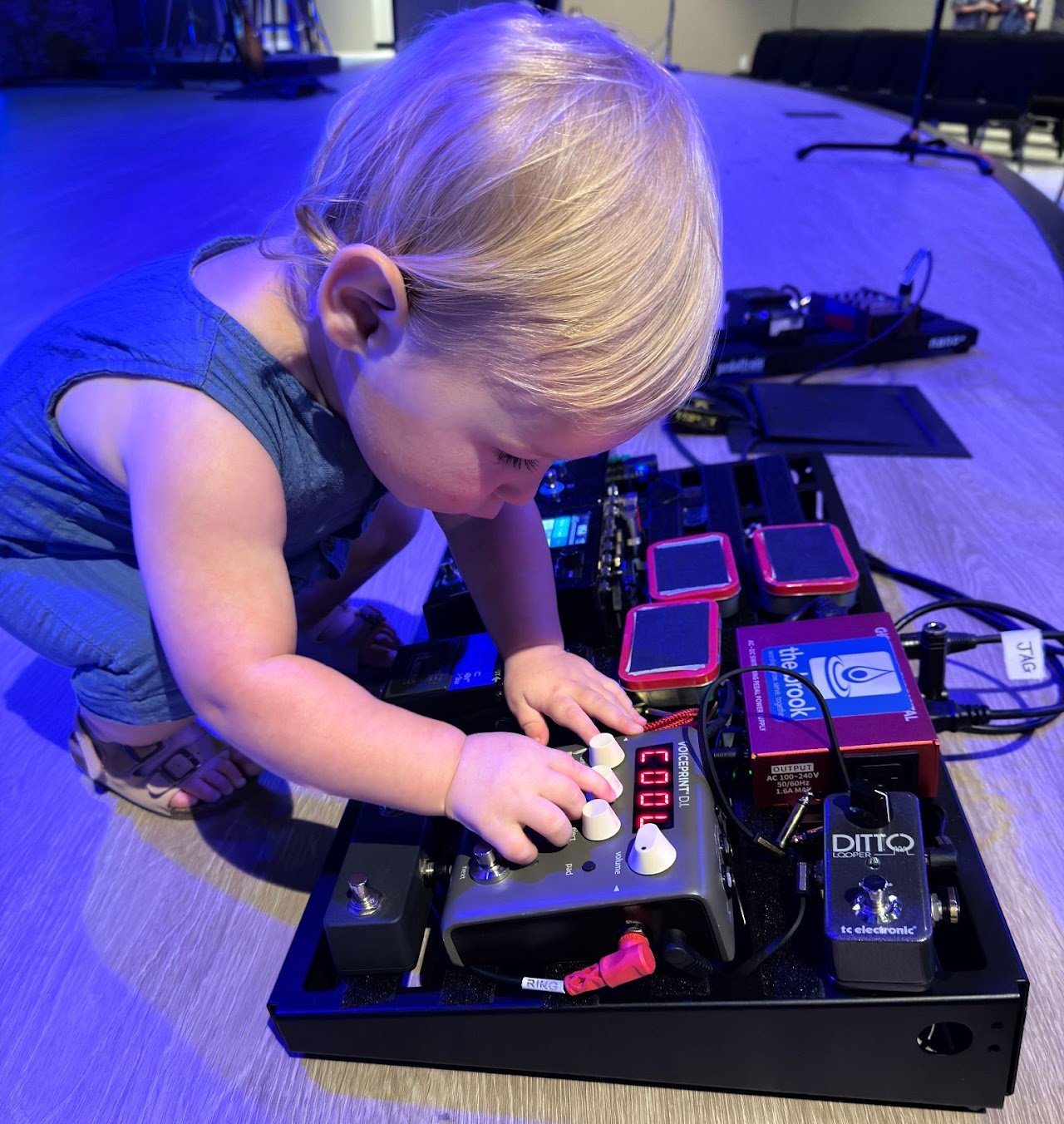 A toddler pressing buttons on a guitar pedalboard on a stage