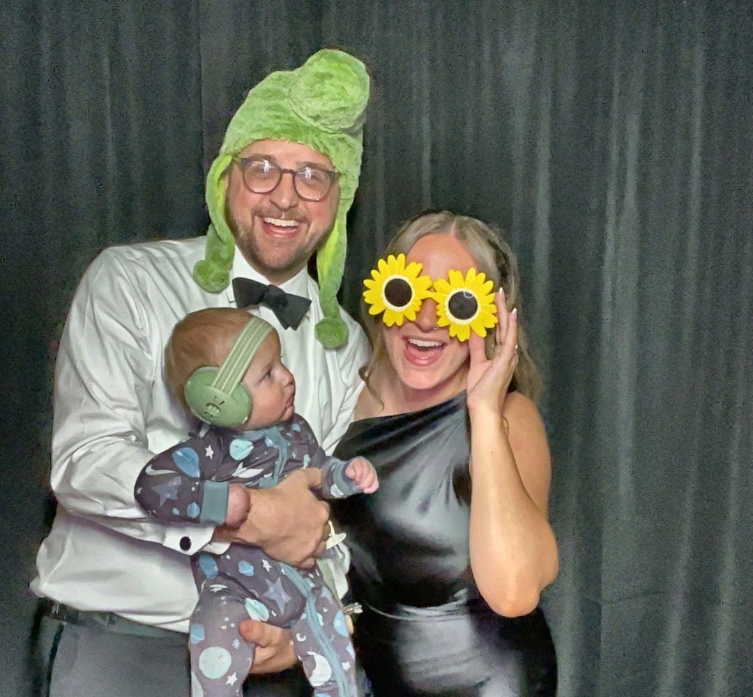 Jon in a frog hat and Katherine in sunflower glasses holding baby Leo in a photo booth