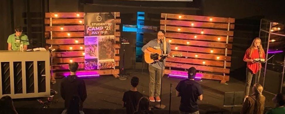 Jon leading worship with students on a stage with a lit wooden pallet backdrop and cross