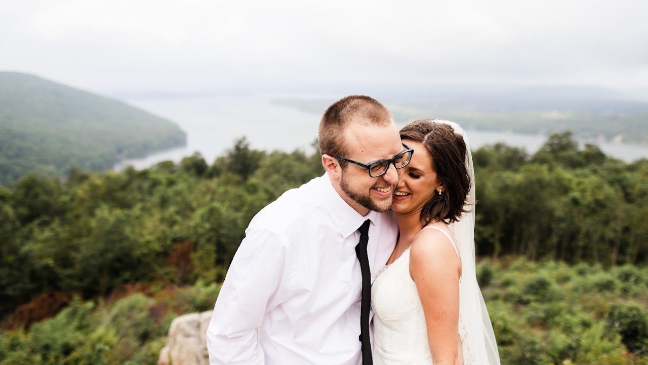 Jon in a white shirt and tie with Katherine in her wedding dress, overlooking a lake and hills